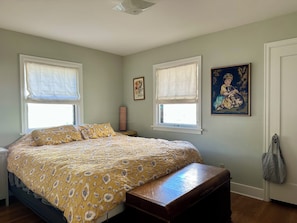Quarto com cama, duas janelas com persianas, porta e um quadro na parede.