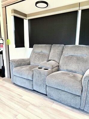 A grey sectional sofa with a remote control on it, positioned against a wall with a black panel and a white ceiling. - Luxury glamping suite near beaches & OWA, POOL! (Foley)