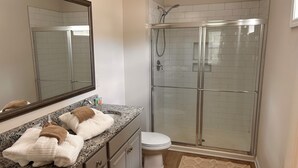 Un baño con encimera de granito, inodoro blanco, ducha con mampara de vidrio y alfombra color beige.