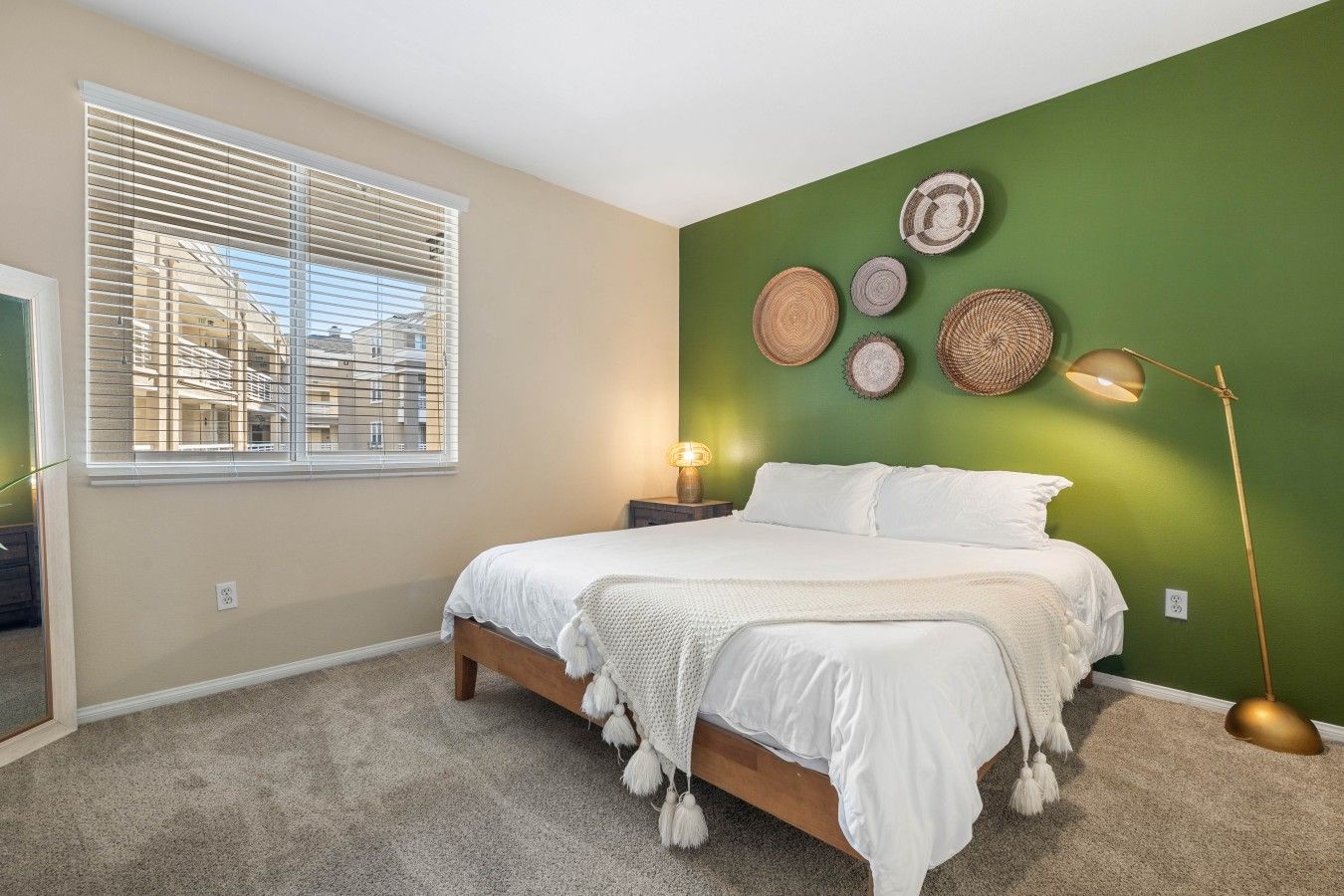 A bedroom with a bed, a window with blinds, a wall with decorative plates, and a floor lamp.