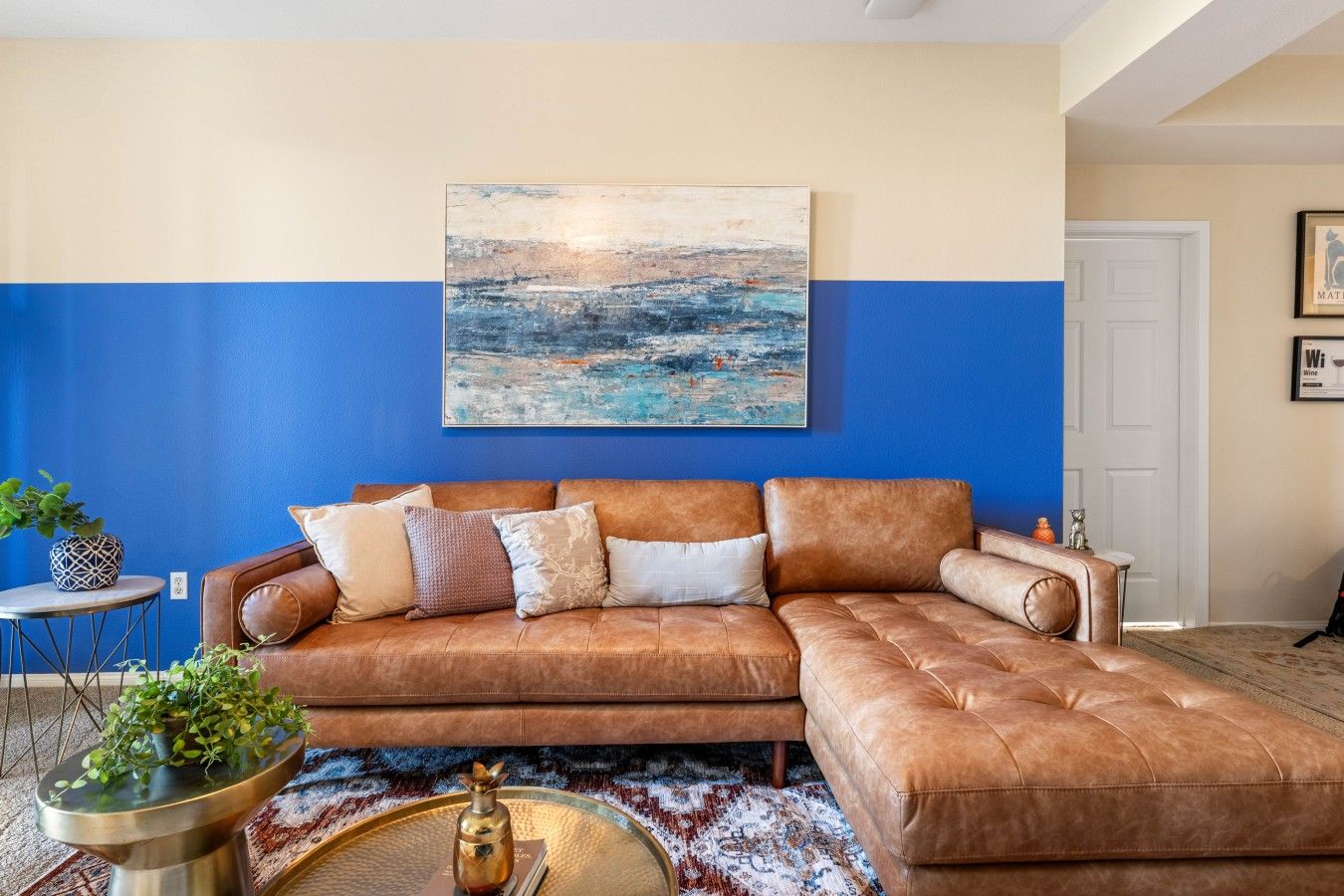 A living room with a brown leather sectional sofa, a blue and beige wall, a large abstract painting, and a small round table with a plant.