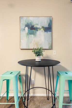 A room with a round table, two stools, a potted plant, and a painting on the wall.
