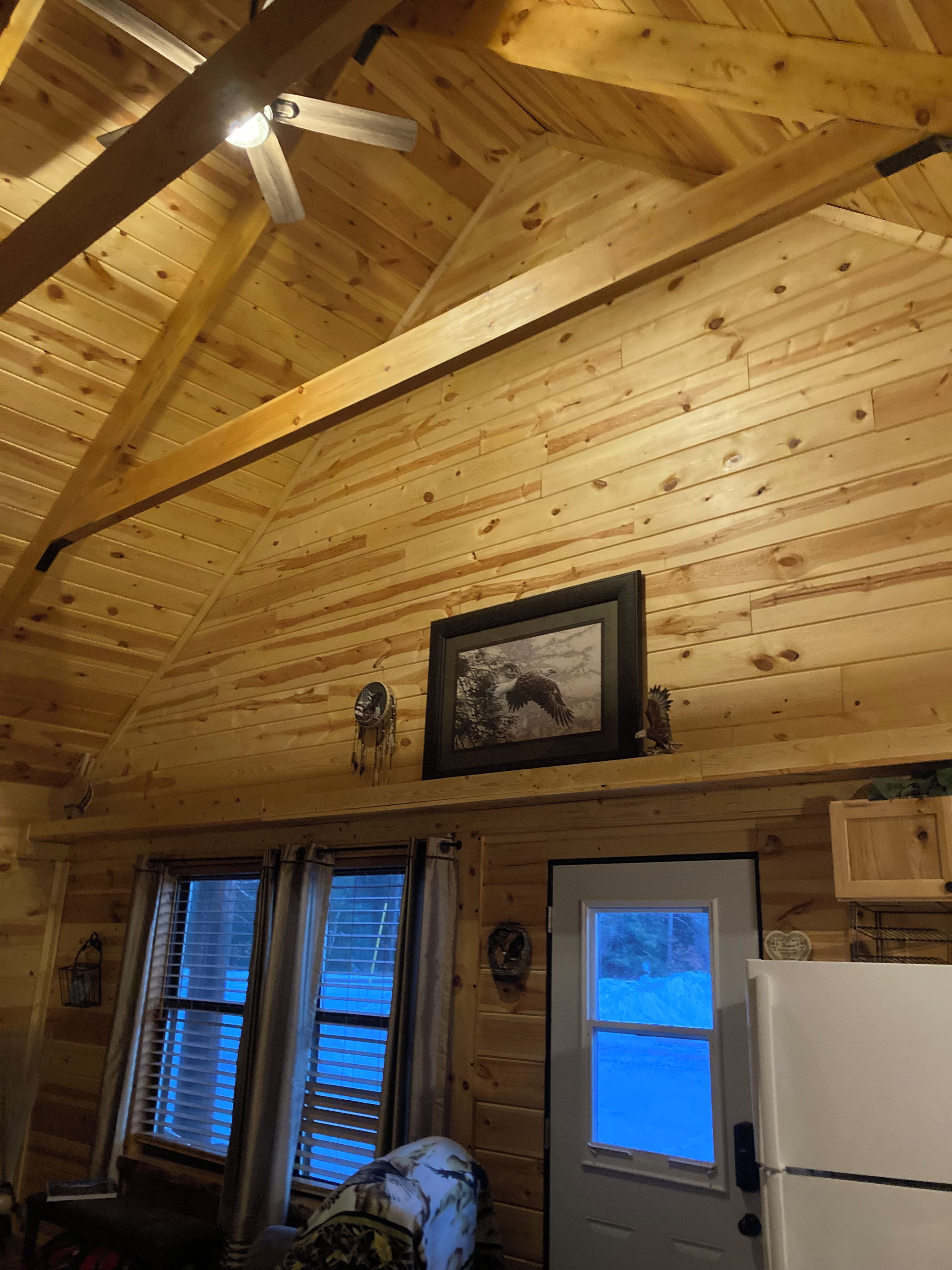 Un interior de madera con un ventilador de techo, dos ventanas con persianas, una puerta y un refrigerador.