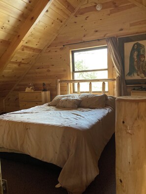 Een slaapkamer in een houten hutje met een enkel bed, een dressoir en een raam met gordijnen.