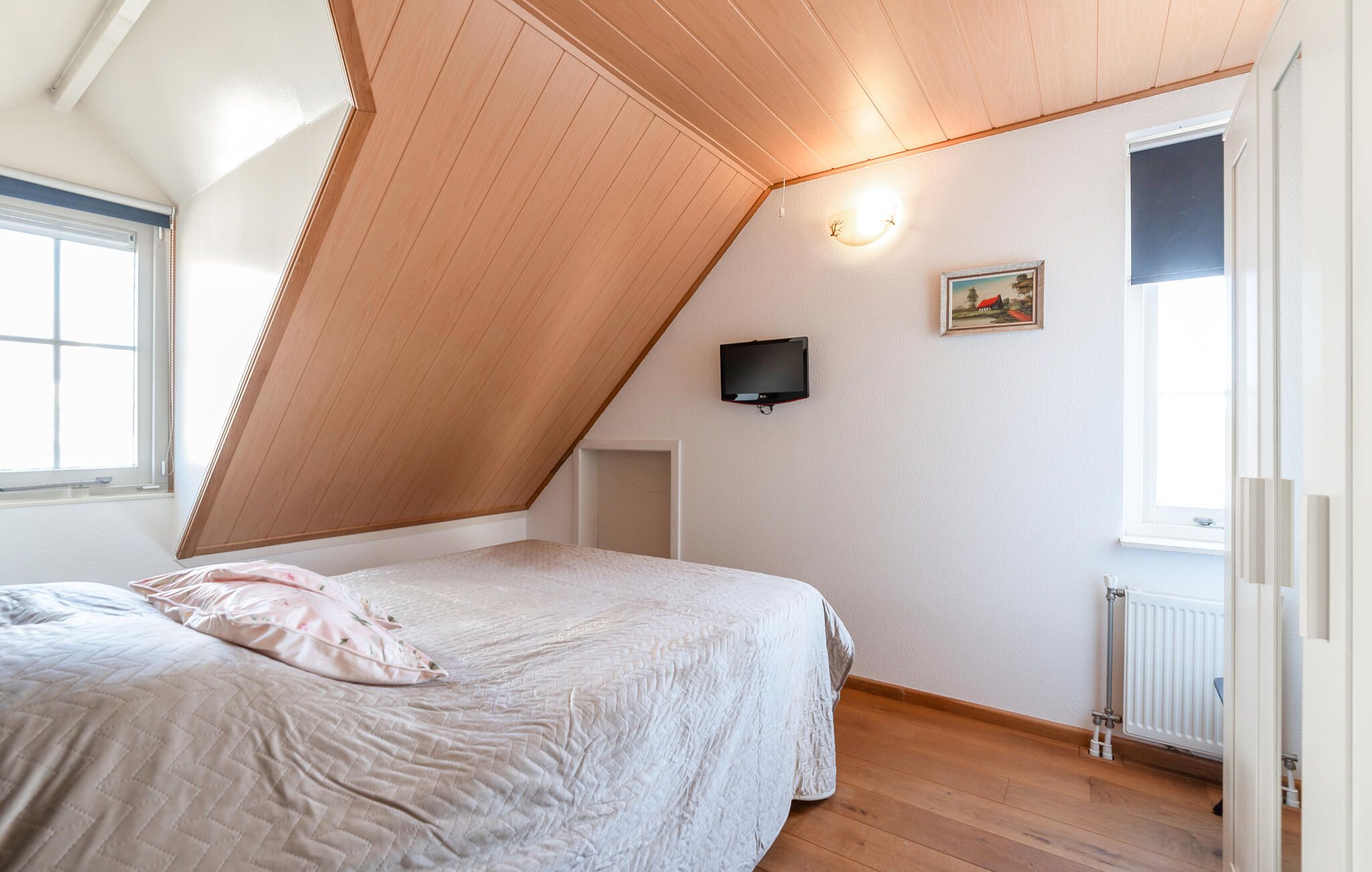 Een slaapkamer met een schuine balkenlaag, een bed met sprei, een aan de muur bevestigde televisie en een radiator.