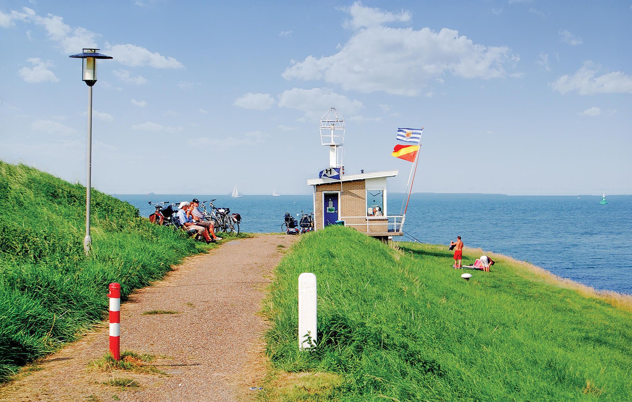 Un sendero costero con una pequeña construcción, una bandera y un faro en la distancia.