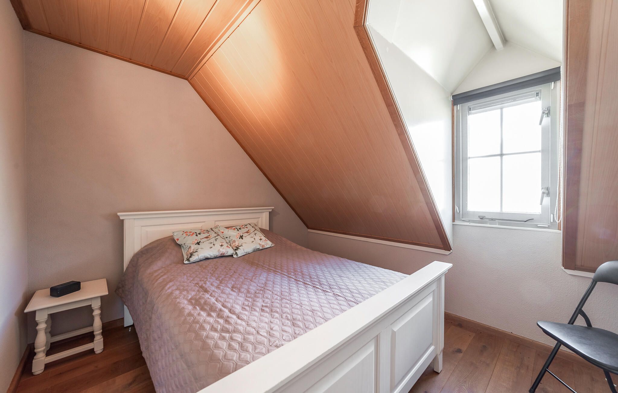 Ein Schlafzimmer mit schrägem Dach, einem Bett mit einem Blumenkissen, einem kleinen Nachttisch und einem Fenster mit Vorhängen.