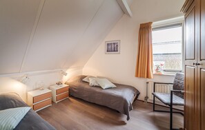 Quarto com cama, mesas de cabeceira, uma cadeira, janela com cortinas e radiador.