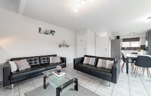 Ein modernes Wohnzimmer mit zwei schwarzen Ledersofas, einem Glastisch und einer im Hintergrund sichtbaren Küchenzeile.