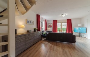 A modern living room with a sofa, a dining table, and a TV. There is a staircase on the left and a window with curtains. - Awesome home in Wemeldinge with sauna (Wemeldinge)