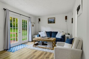 A cozy living room with a sofa, a coffee table, and a chair. There is a sliding glass door leading to a garden.