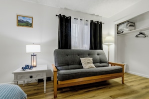 A room with a grey sofa, a small bedside table with a lamp, a framed picture on the wall, and a window with curtains.