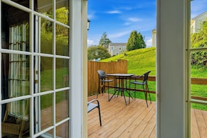 An open door leading to a wooden deck with a table and chairs, overlooking a grassy area and a hill with a house in the background.