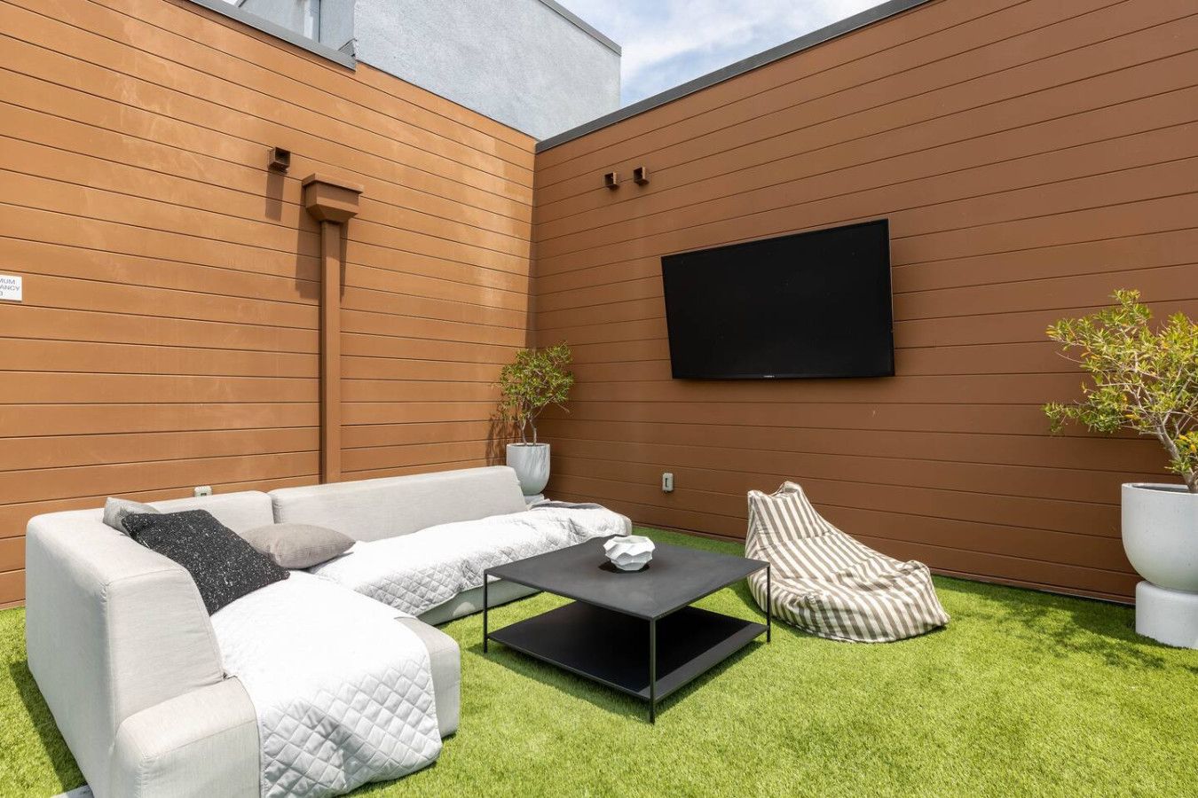 A modern outdoor seating area with a sofa, armchair, and coffee table, a flat-screen TV mounted on the wall, and two potted plants.
