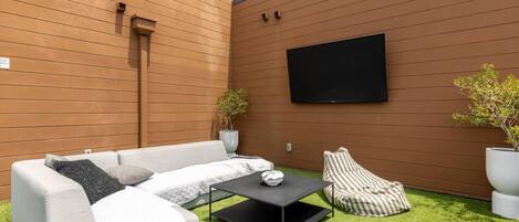 A modern outdoor seating area with a sofa, armchair, and coffee table, a flat-screen TV mounted on the wall, and two potted plants.