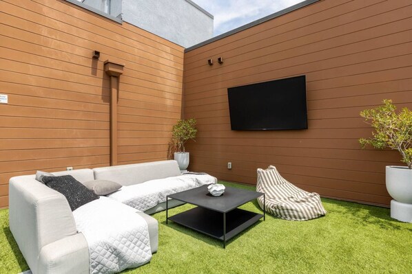 A modern outdoor seating area with a sofa, armchair, and coffee table, a flat-screen TV mounted on the wall, and two potted plants.