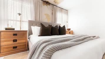 Un lit bien fait, recouvert d’une couverture tricotée grise, une table de chevet en bois avec une lampe, et une autre table de chevet avec un livre.