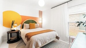 Ett hotellrum med en säng, ett sängbord, en lampa och ett fönster med persienner.