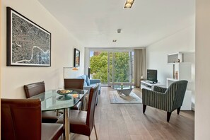 Ein modernes Wohnzimmer mit einem Glastisch, einer Couch, einem Fernseher und einem Balkon mit Blick auf Bäume.