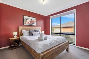 A bedroom with a large bed, a nightstand, a lamp, a framed picture on the wall, and a window with a view of a building. - Blue Vista Retreat - Apollo Bay (Apollo Bay)