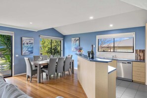 A modern kitchen with a central island, a dining area with a table and chairs, and a view of the outdoors through large windows. - Blue Vista Retreat - Apollo Bay (Apollo Bay)