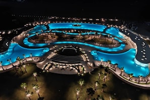 A large, well-lit swimming pool complex with multiple sections and surrounding palm trees. - Sonoran Star Beachfront 2BR - Ocean & Pool Views · Jacuzzi · Beach Access (Puerto Peñasco)
