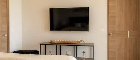 Quarto com cama, televisão presa à parede, mesa de cabeceira com objetos decorativos e guarda-roupa.
