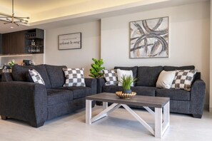 A modern living room with a grey sofa set, a coffee table, and decorative pillows. There is a kitchen area in the background with a wine rack and a sign that reads 'welcome to ours.' - Sonoran Star Beachfront 2BR - Ocean & Pool Views · Jacuzzi · Beach Access (Puerto Peñasco)