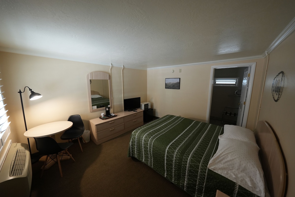 Habitación de hotel con cama, escritorio, silla, televisor y ventana con persianas.
