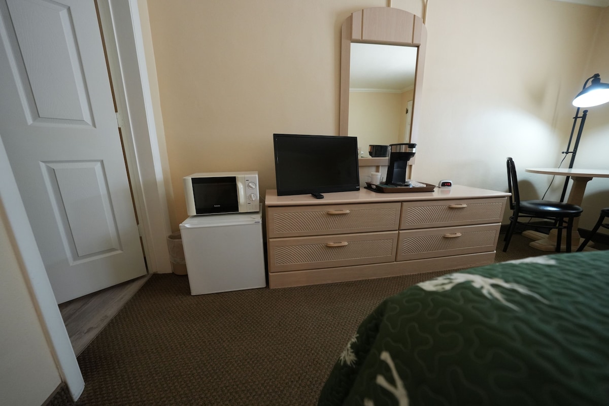 Una habitación de hotel con cama, escritorio con lámpara, microondas, televisión y cómoda.