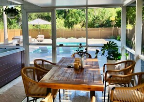 Eine überdachte Terrasse mit Holztisch und Korbstühlen, mit Blick auf einen Swimmingpool.