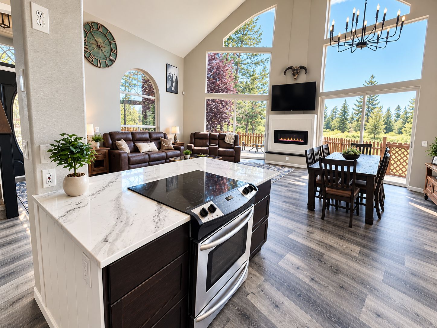 A spacious living area with a large island, dark wood flooring, a fireplace, and a view of trees through large windows.
