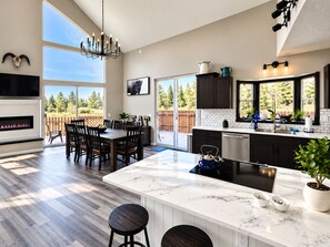 A modern kitchen with a large island, a dining area with a fireplace, and a view of a wooden deck. - River Front Getaway! Hot tub & Pool Table. Great location to Sunriver and Bend (Bend)