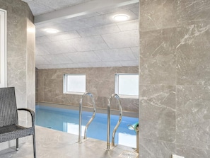 Una piscina interior con agua cristalina, dos escaleras metálicas y una silla.
