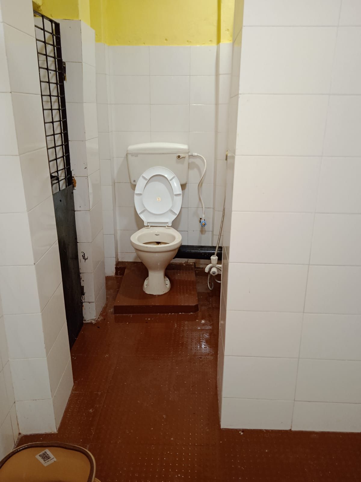 A small, narrow bathroom with a toilet, a visible pipe, and a metal grate on the left wall.
