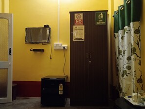 Habitación con pared amarilla, puerta cerrada con letrero de 'NO FUMAR', señalización de cesto de basura, un refrigerador pequeño y cortina con estampado floral.