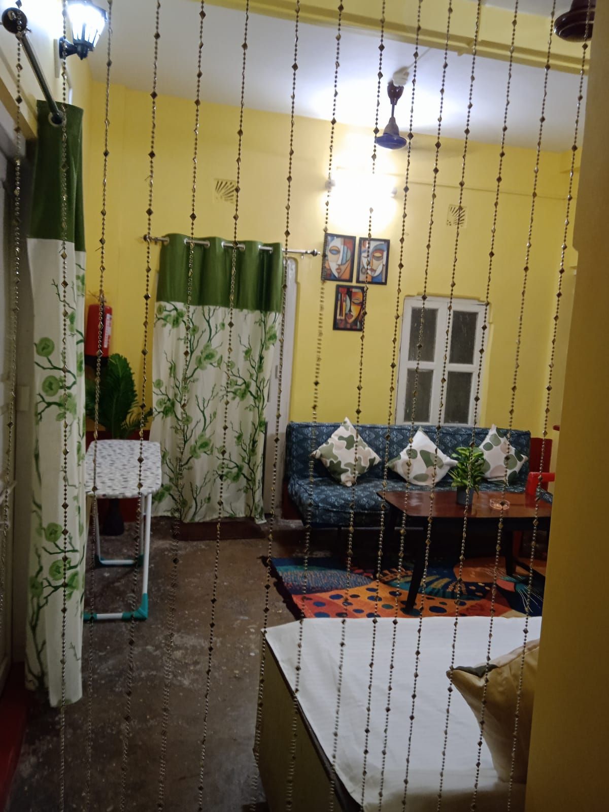 A living room with a sofa, a small table, and a chair. There is a window with a curtain, and a plant on the table.