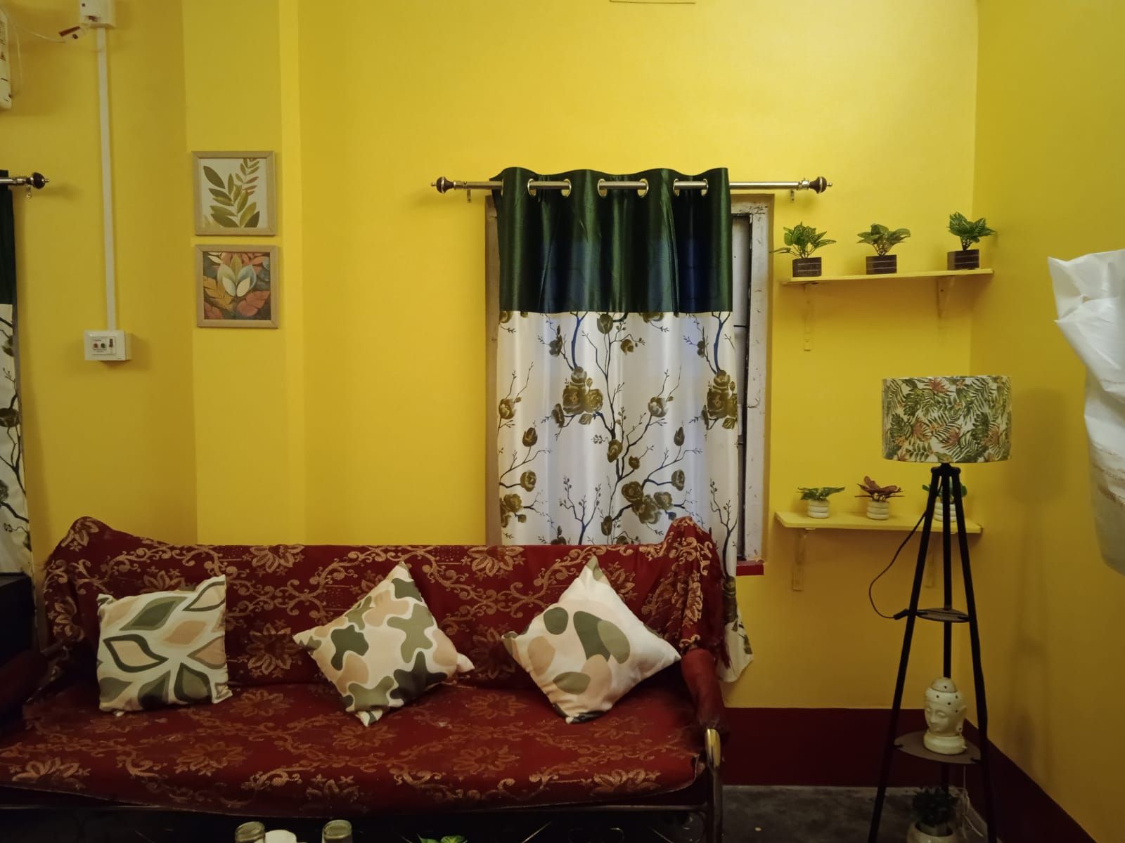 Una habitación con sofá rojo, cojines estampados, cortina verde, una estantería pequeña con plantas y una lámpara de pie.