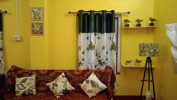 Una habitación con sofá rojo, cojines estampados, cortina verde, una estantería pequeña con plantas y una lámpara de pie.