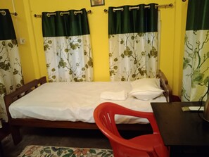 Una cama individual con sábana blanca, una silla de plástico roja y una mesita con un distribuidor de corriente.