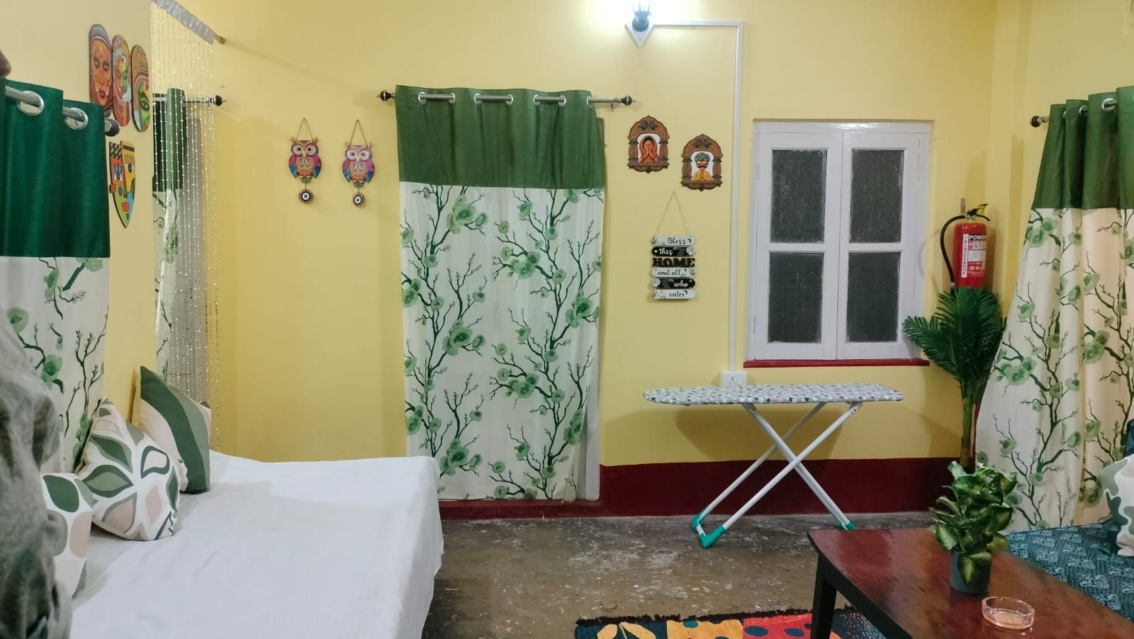Una habitación con cama, una mesita, un ventanal y una cortina con estampado de árboles.