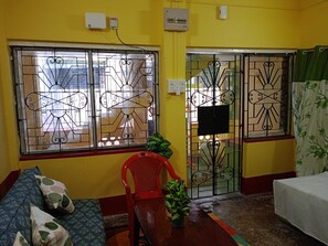 Una habitación con paredes amarillas, una cama, una silla roja, un sofá con cojines estampados, una planta en maceta y ventanas con barrotes.
