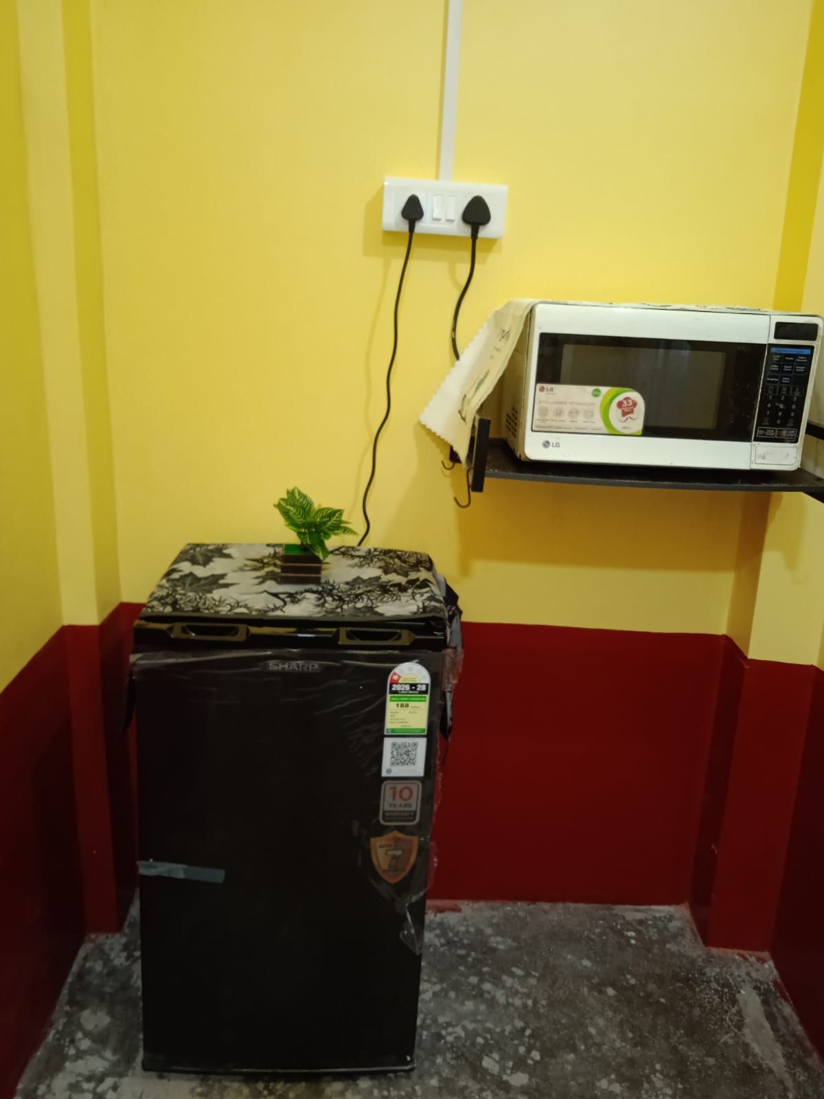 Un refrigerador negro con una pegatina, un microondas blanco sobre una repisa y un enchufe en la pared.