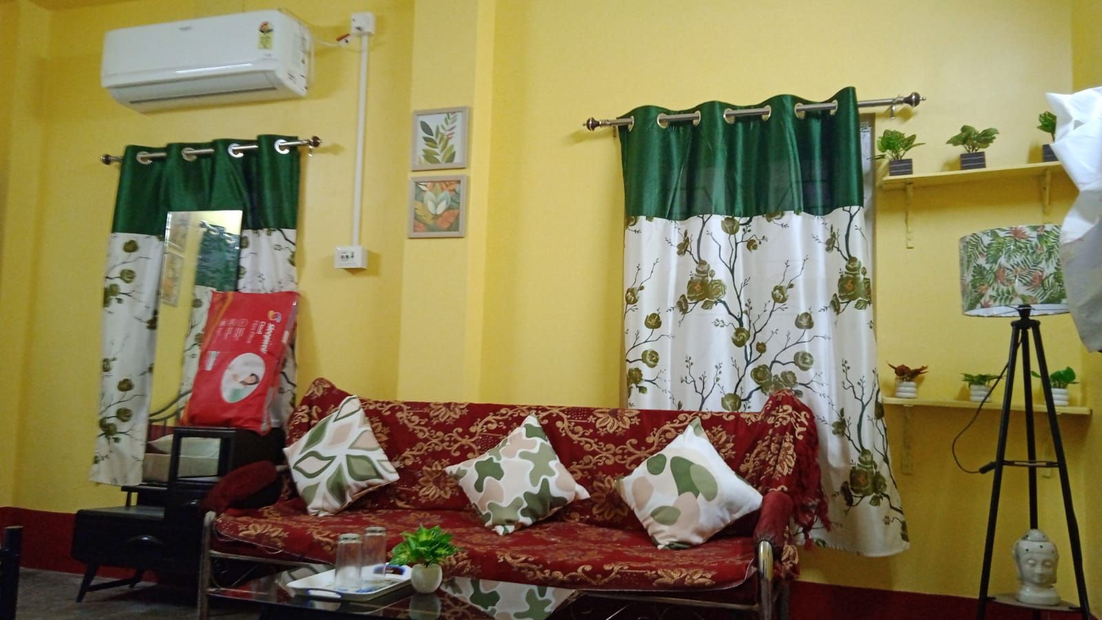Una sala de estar con un sofá rojo, cortinas verdes y blancas, una mesita con una planta y un aire acondicionado instalado en la pared.