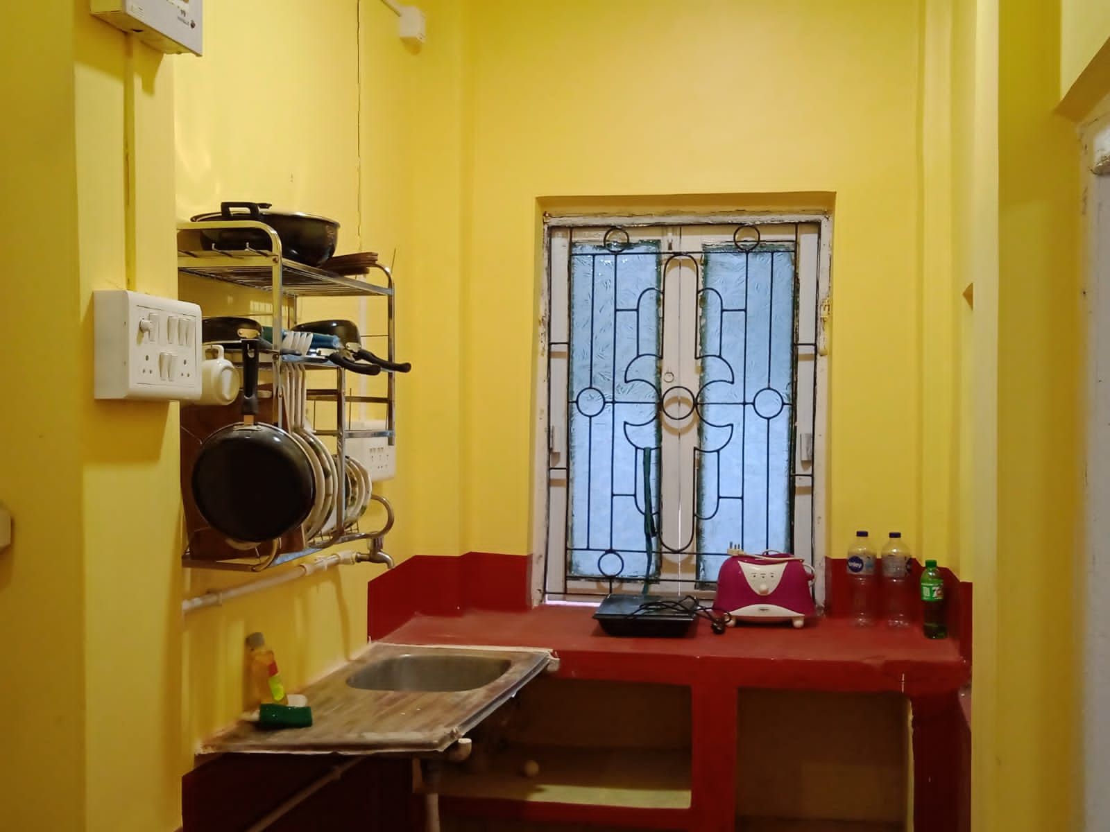 Eine kleine Küche mit Spüle, einem Fenster mit Metallgitter und einem Regal mit Geschirr und Toaster.