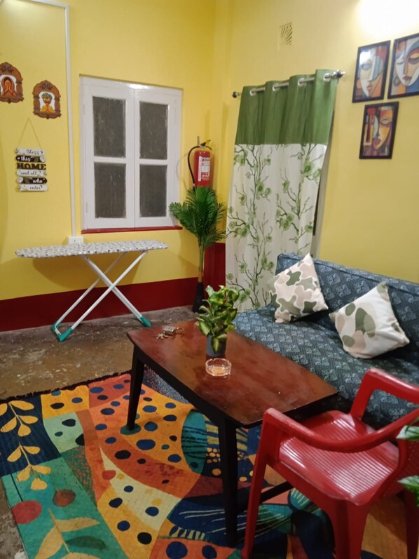 Una habitación con sofá, una silla roja, alfombra estampada, ventana con cortinas y extintor de incendios fijado a la pared.