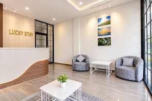 Un área de recepción con un mostrador de madera, dos sillones grises, una pequeña mesa blanca y una planta en maceta.