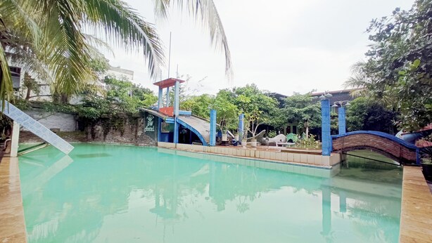 A swimming pool with a bridge, surrounded by greenery and a building with a sign. - Collection O near Mall Ciputra (Pekanbaru)