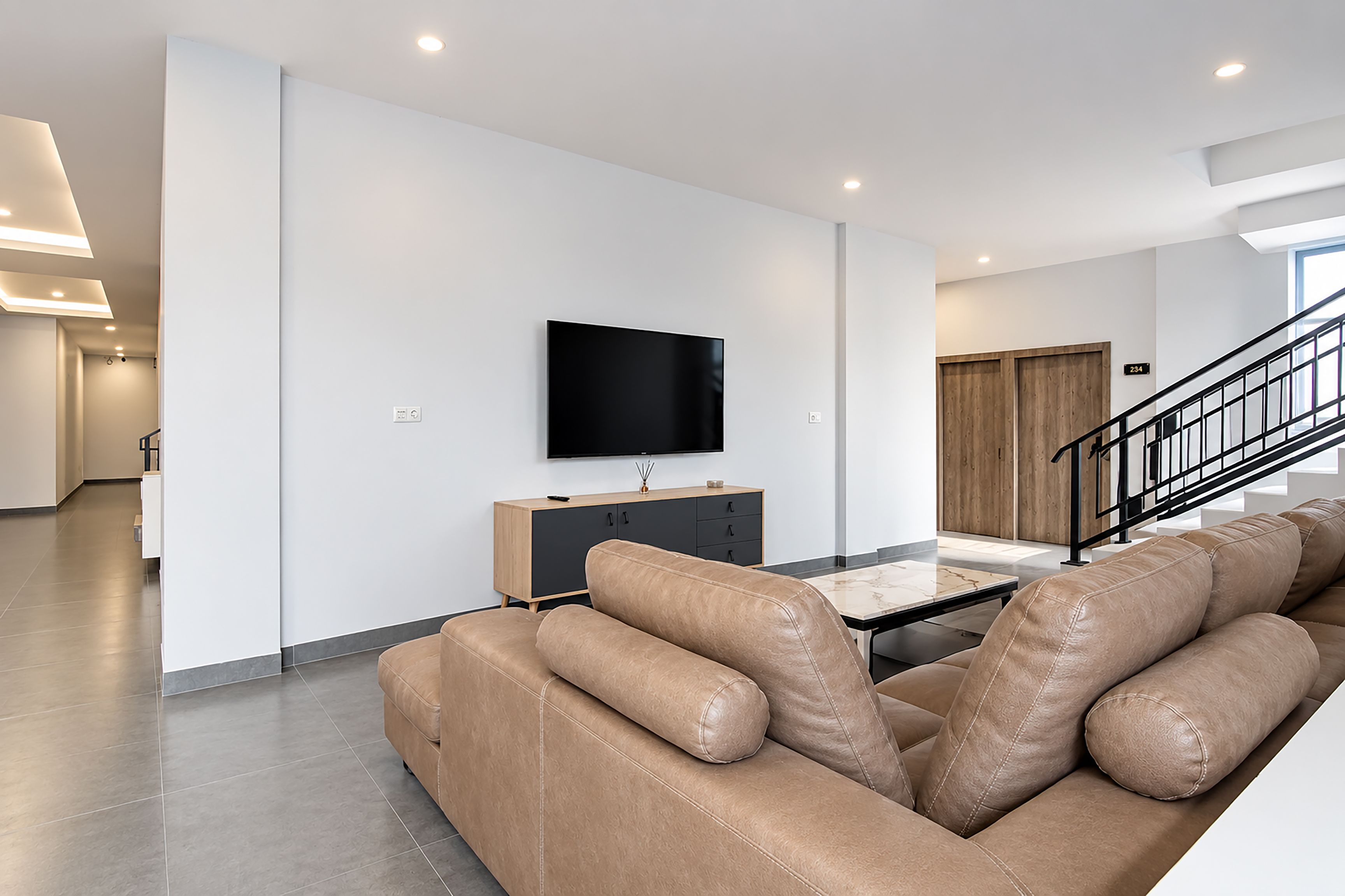 A modern living room with a leather sofa, a flat-screen TV mounted on the wall, and a staircase leading to another level.