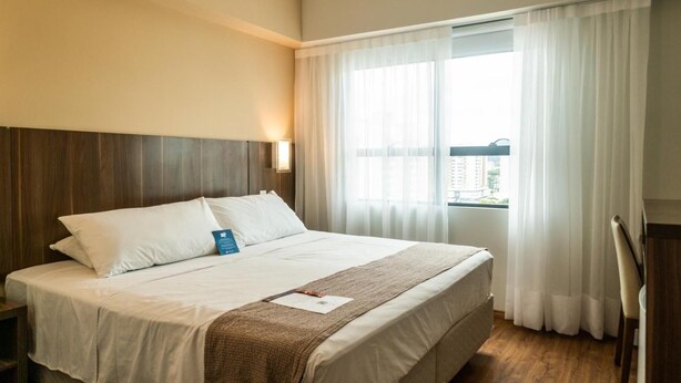 Quarto de hotel com uma cama, mesa de cabeceira, janela com cortinas e vista para edifícios.